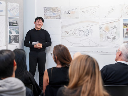 A student wearing black sweater and pants, standing and smiling in front of boards with drawings and illustrations, and facing a few rows of people
