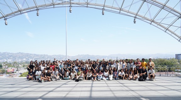 A group of about fifty people, posed in rows with gray asphant in front and a blue sky and mountains in the background