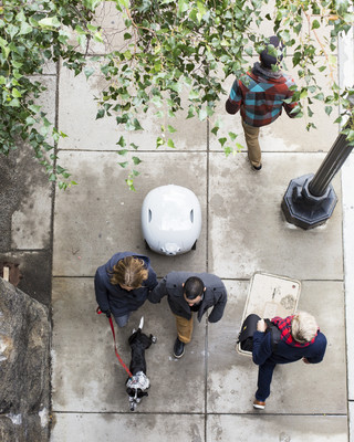 A bird's eye view of Gita following a couple walking with a dog in the street. Gita is a white, droid-like personal autonomous assistant that is able to carry luggage while it follows its owner on its own.