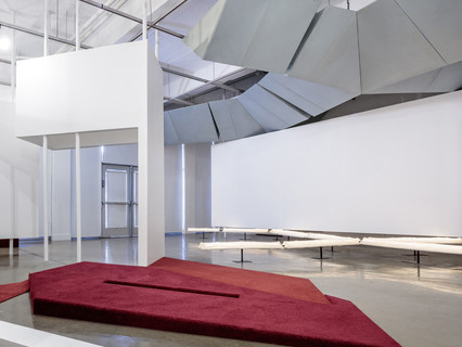 Image of an exhibition featuring red carpet and metal duct-like sculpture hanging from the ceiling