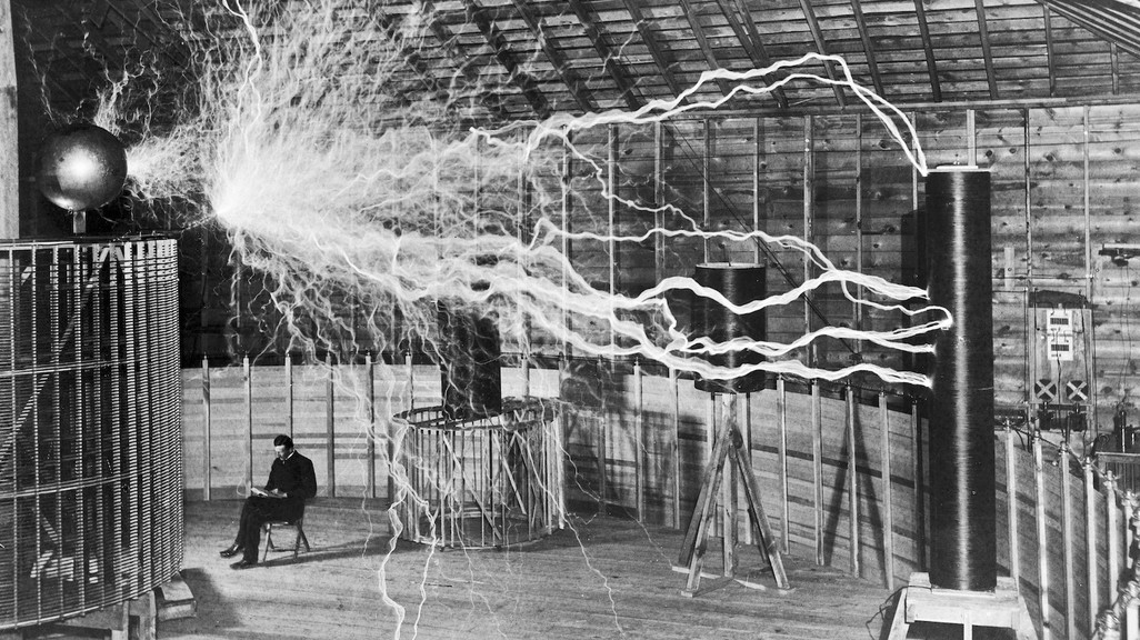 A black-and-white photograph of an inventor sitting beneath an electromagnetic experiment, showing a man seated beneath a large explosion of electricity between two metal conductors