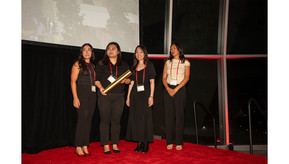 Four women stand in a row on a red carpet, each dressed in black outfits, receiving an award in front of a projector screen showing their work