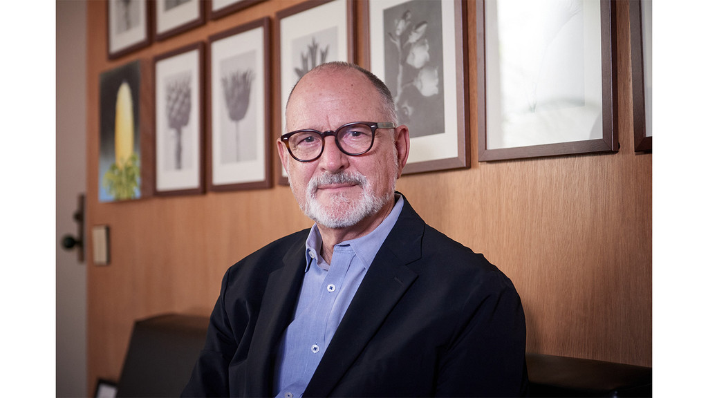 A man with round framed glasses and a blue long sleeved shirt with black suit jacket, sitting and smiling in front of a wood patterened wall with framed art