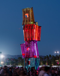 A photograph of a tall structure made of different, red-colored panels and screens, placed on a large open lawn area with a blue sky and people standing around the structure