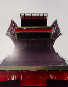 A photograph of a tall structure made of different, red-colored panels and screens, placed on a large open lawn area with a blue sky and people standing around the structure