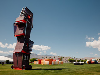 A photograph of a tall structure made of different, red-colored panels and screens, placed on a large open lawn area with a blue sky and people standing around the structure