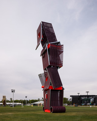 A photograph of a tall structure made of different, red-colored panels and screens, placed on a large open lawn area with a blue sky and people standing around the structure