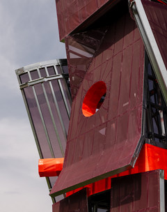 A photograph of a tall structure made of different, red-colored panels and screens, placed on a large open lawn area with a blue sky and people standing around the structure