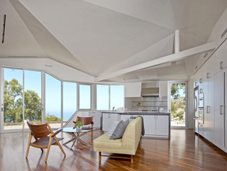 A photograph showing a big, open living room and kitchen, with tan wood floors, a geometrical white ceiling, and some chairs and a couch in the center