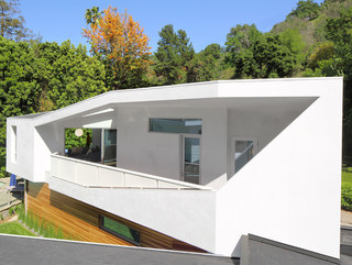 A photograph showing the outside of a geometric, white house, with a large opening on a long exterior wall, showing windows and rooms inside, and with green trees in the background