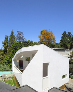 A photograph showing the outside of a geometric, white house, with a large opening on a long exterior wall, showing windows and rooms inside, and with green trees in the background
