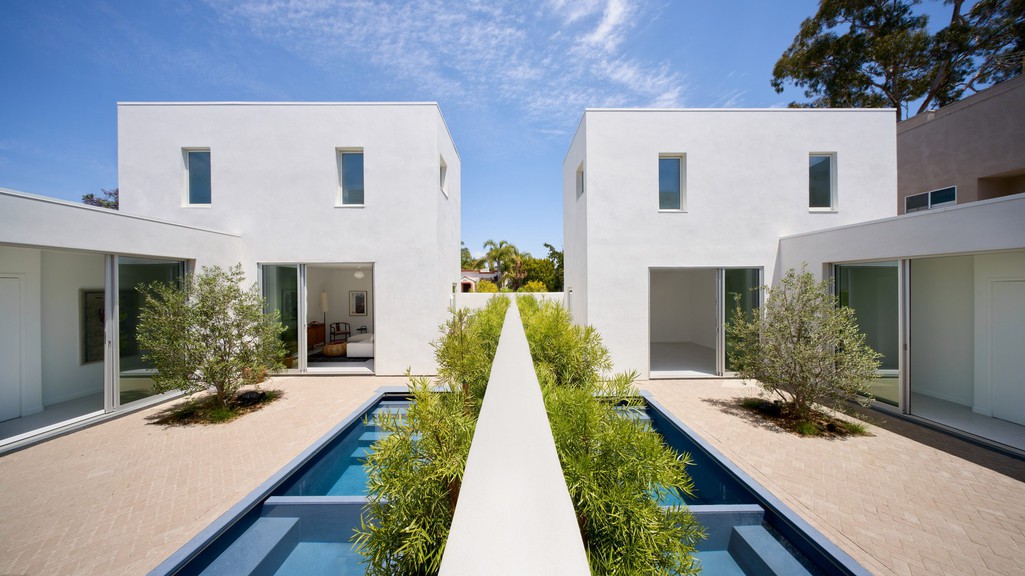 A wide photograph showing two twin houses, each rectangular and made of white stucco, with a pool and brick courtyard in the middle and blue sky in the background