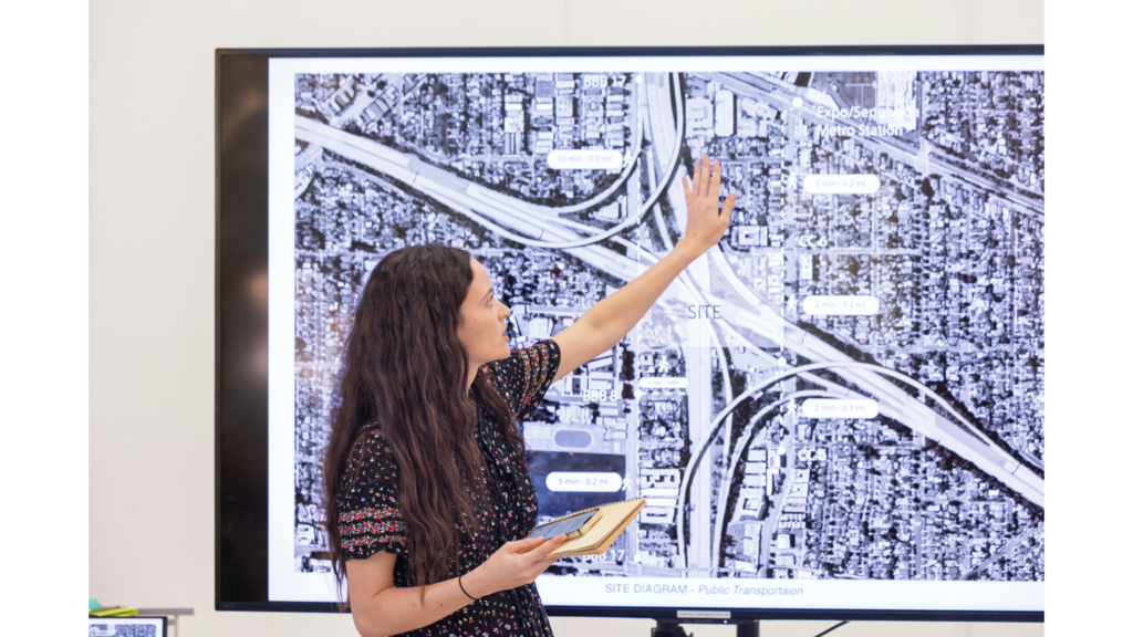 A woman is standing in front of a TV screen showing a map of a Los Angeles freeway cross-over, and is gesturing with her hands to point out parts of the surrounding neighborhood