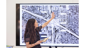 A woman is standing in front of a TV screen showing a map of a Los Angeles freeway cross-over, and is gesturing with her hands to point out parts of the surrounding neighborhood