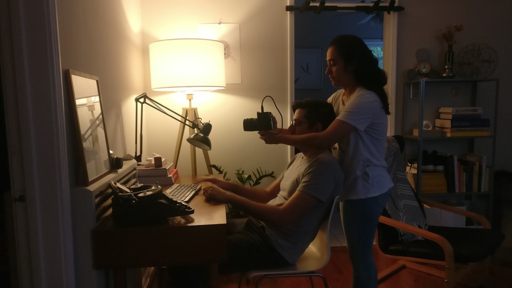 Image of a woman holding a camera over a man seated in front of a desktop computer